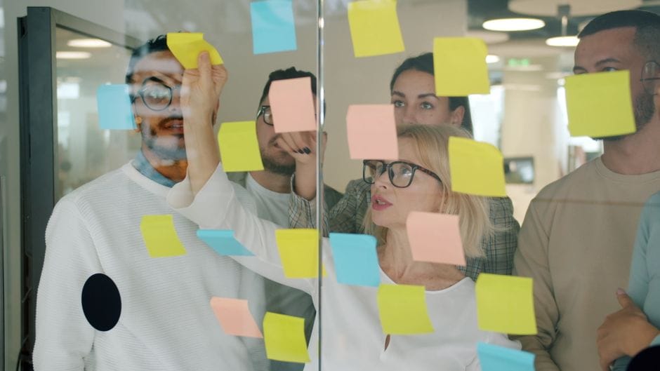 Une équipe aux profils variés se réunit pour un brainstorming à l'aide de post-it colorés sur une vitre dans un bureau moderne.