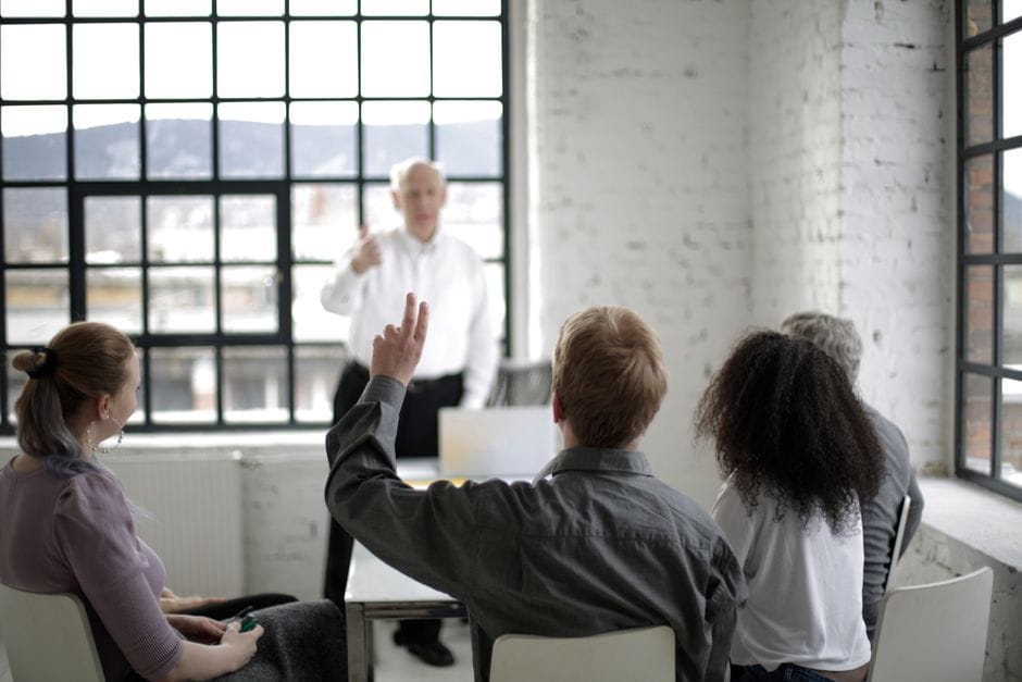 Groupe d'adultes en discussion, avec une personne levant la main dans un espace de bureau lumineux.