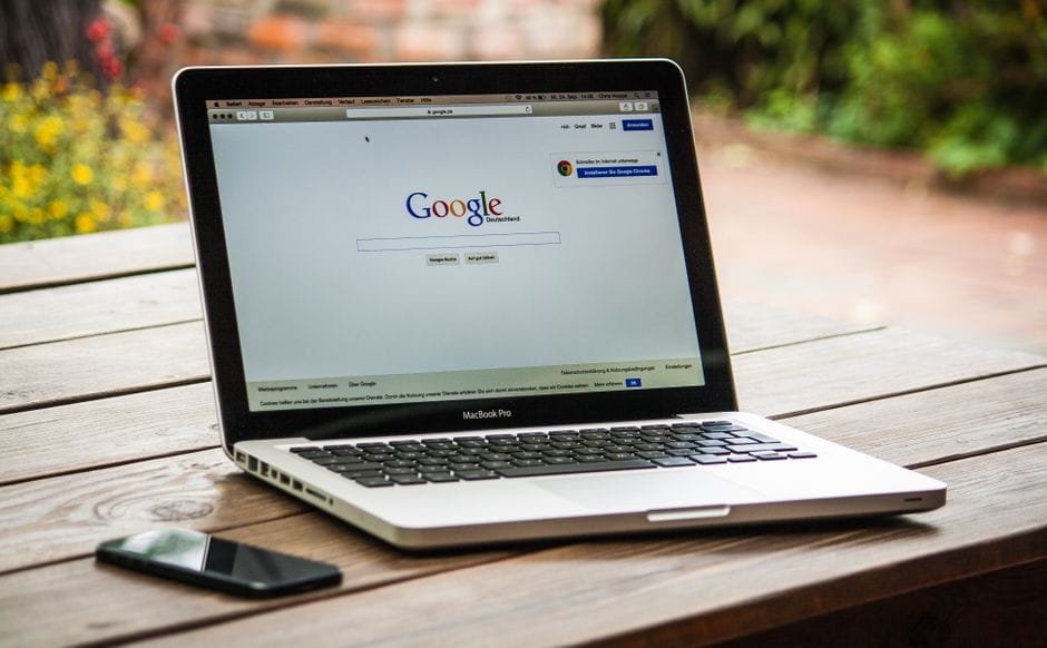 Un MacBook Pro affichant la recherche Google sur une table en bois à l'extérieur, à côté d'un smartphone.