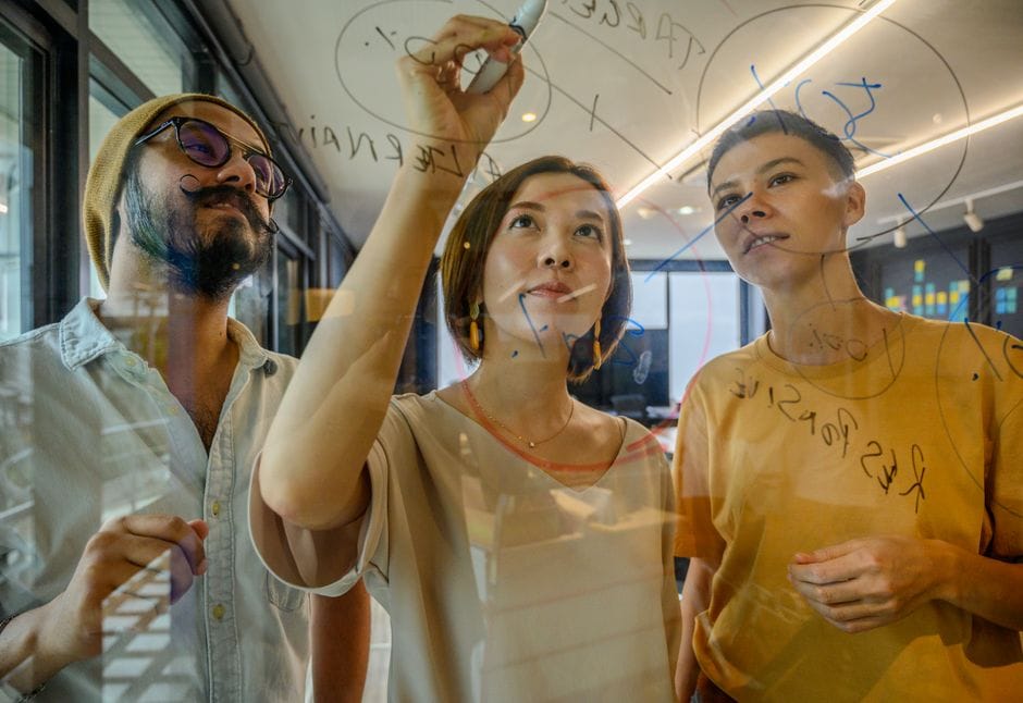 Groupe de professionnels divers collaborant sur un tableau en verre dans un cadre de bureau moderne.